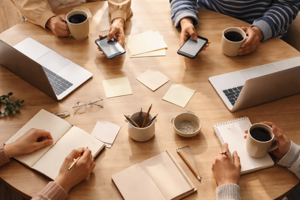 Mission/values-billede: Et mere abstrakt, symbolsk billede af forskellige hænder, der rækker ind over et bord med spredte sedler og laptops, som symboliserer deling af historier, fællesskab og tryghed omkring det pinlige.